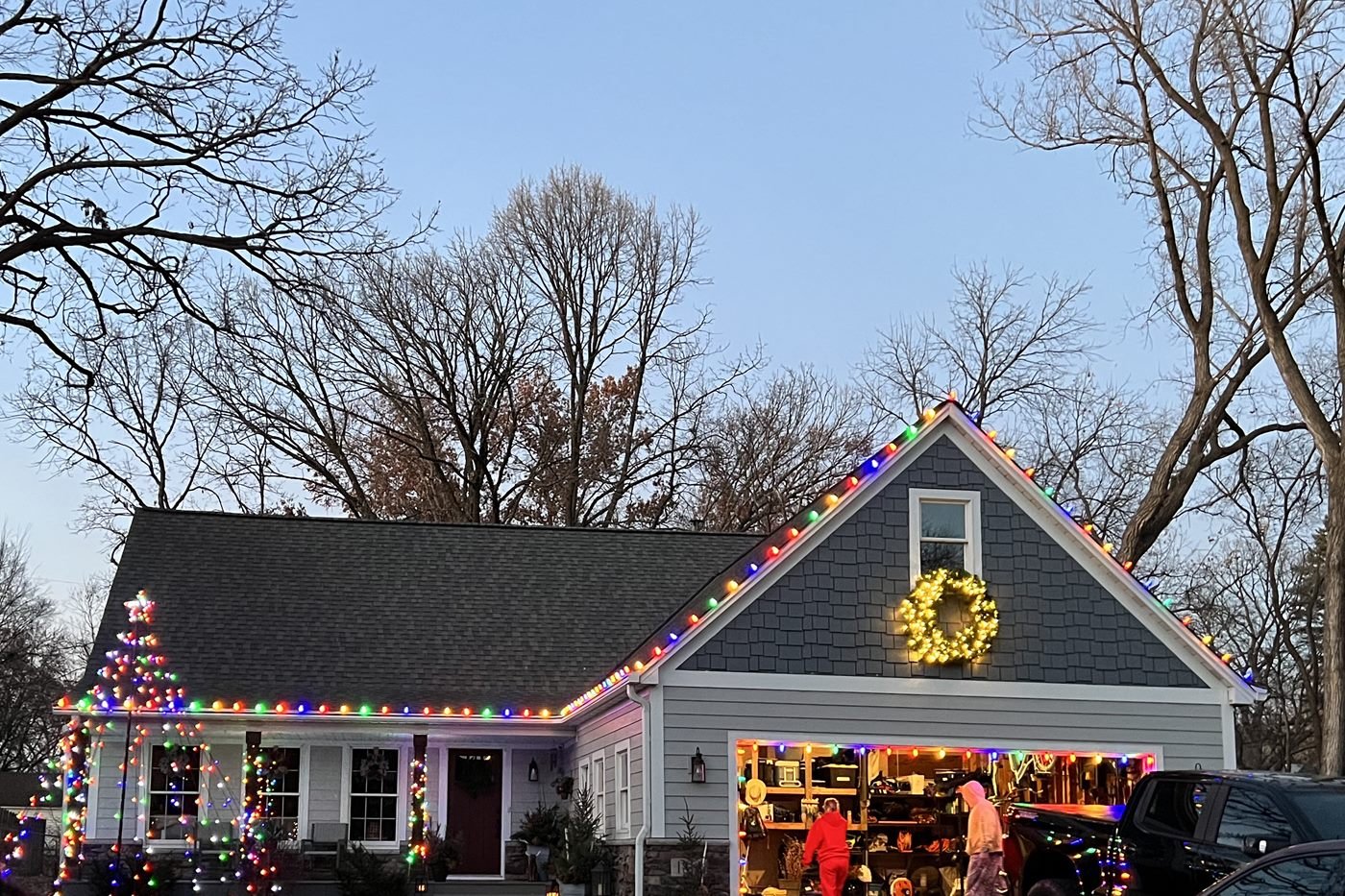A home with multicolour holiday lights along the roofline and peak, a lit wreath on the gable and wrapped trees in the front yard